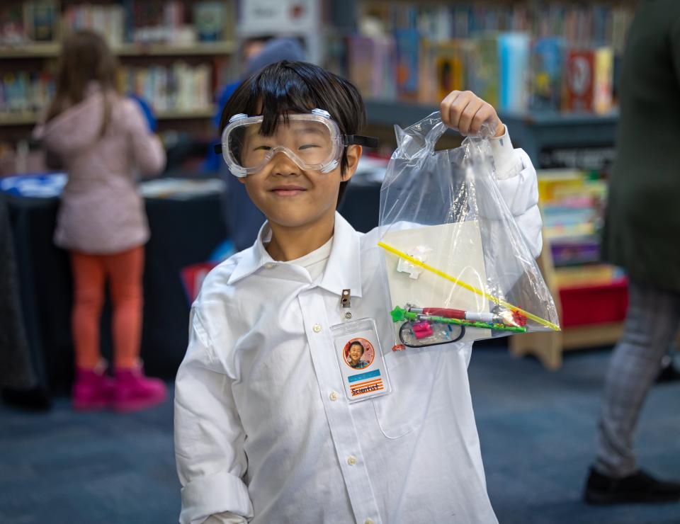 Elementary School student dressed in a lab coat and goggles stands in a library, holding up a plastic bag of school supplies.